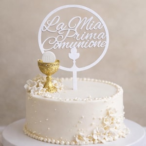 Può includere: Una torta bianca decorata con glassa bianca, perle e decorazioni floreali. Un calice dorato con un'ostia è posto sopra la torta, e un topper bianco recita "La Mia Prima Comunione".