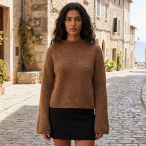 May include: A woman wearing a brown knit sweater with long sleeves and a black skirt. The sweater has a crew neck and a slightly flared sleeve. The woman is standing on a cobblestone street in front of stone buildings.