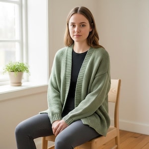 May include: A woman wearing a sage green cardigan over a black top and grey trousers, seated on a wooden chair. The cardigan has a ribbed texture and a relaxed fit. A window and a potted plant are in the background.