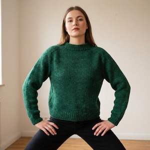 May include: A green knit sweater with a crew neck and long sleeves. The sweater is a solid color and appears to be made of a soft, textured yarn. The person is wearing black pants.