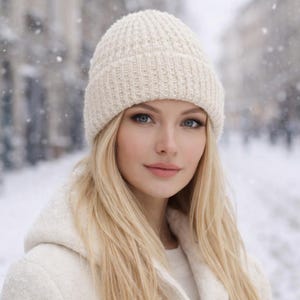 Op de afbeelding: Een vrouw met blond haar draagt een crèmekleurige gebreide muts en een bijpassende jas. De achtergrond is een besneeuwde straat met vallende sneeuwvlokken. De vrouw heeft blauwe ogen en kijkt in de camera.