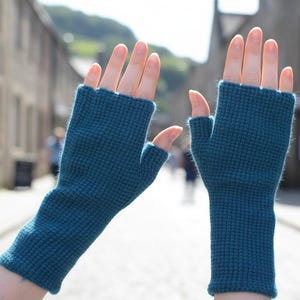 Puede incluir: Guantes sin dedos de color turquesa con un patrón de punto texturizado. Los guantes se muestran en una persona con las manos levantadas, mostrando el diseño y el ajuste. Los guantes son de color turquesa liso y parecen estar hechos de un material cálido.