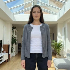 Op de afbeelding: Een grijze cardigan met kabelgebreide details aan de voorkant en mouwen. Gedragen over een witte t-shirt en zwarte jeans. De persoon staat in een lichte kamer met een modern interieur.