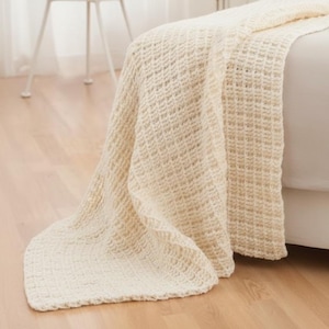 May include: Cream-coloured woven blanket with a waffle-like pattern draped over a bed. The blanket hangs onto a wooden floor. A white chair is in the background. The blanket is a neutral colour.