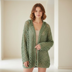 Puede incluir: Un cárdigan con capucha verde claro y abierto por delante. El cárdigan está hecho de un material de punto suelto, con un diseño de tejido abierto y texturizado. Las mangas son largas y el cárdigan llega hasta la mitad del muslo. La prenda se lleva sobre una camiseta de color neutro.