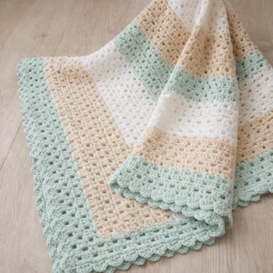 May include: A handmade crochet blanket in soft pastel colors. The blanket features alternating stripes of white, light beige, and mint green. The edges are finished with a scalloped border, adding a delicate touch to the cozy, textured design.