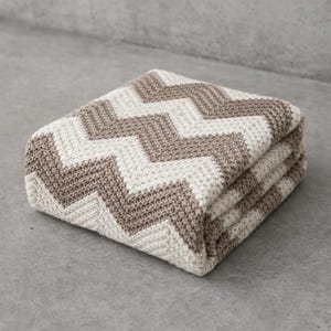 Puede incluir: Manta plegada con un patrón de chevrón en crema y topo. La manta está hecha de una tela de punto texturizada y está doblada cuidadosamente, mostrando el diseño del patrón. La manta es de color neutro y sería adecuada para una variedad de estilos de decoración del hogar.
