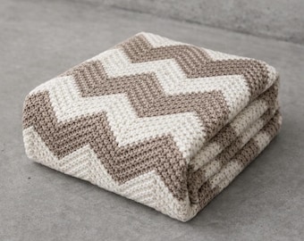 May include: Folded blanket with a chevron pattern in cream and taupe. The blanket is made of a textured knit fabric and is folded neatly, showing the pattern's design. The blanket is a neutral color and would be suitable for a variety of home decor styles.
