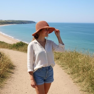 Könnte beinhalten: Eine Frau trägt einen hellbraunen Sonnenhut aus Stroh, ein weißes Hemd mit Knöpfen und Jeansshorts. Sie steht auf einem Feldweg, der zu einem Strand und Ozean unter blauem Himmel führt. Der Hut spendet Schatten.