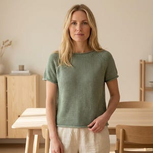 Puede incluir: Una camiseta de punto verde salvia de manga corta con textura acanalada. La camiseta tiene cuello redondo y un ajuste relajado. La modelo lleva pantalones beige. El fondo incluye una mesa de madera, sillas y un armario.