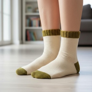 Può includere: Un paio di calzini color crema con tallone, punta e polsini verde oliva. I calzini a maglia a coste sono indossati all'interno, con uno sfondo sfocato di un soggiorno.
