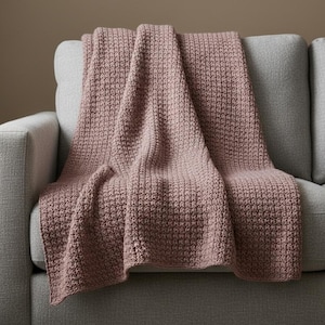 Może przedstawiać: Pierzasta różowa dzianinowa narzuta na szarej sofie. Kocyk ma teksturowany, tkany wzór i jest złożony, tworząc miękkie, płynne linie. Sofa ma neutralny kolor i prosty design.