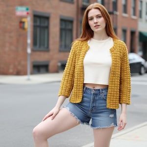 May include: A woman wearing a mustard yellow textured cardigan over a white crop top and denim shorts. The cardigan has a unique woven pattern. The outfit is casual and suitable for warm weather.