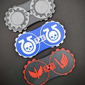 May include: Three colourful, circular game tokens with gear-like edges. One is grey with the number 43, another is blue with a skull and the number 950, and the third is red and black with a winged sword and the number 95.