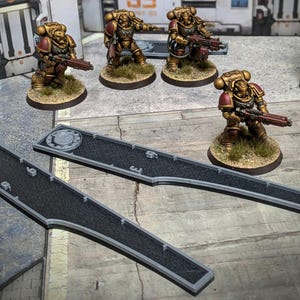 Op de afbeelding: Miniatuurfiguren van gouden en rode gepantserde soldaten met geweren, op getextureerde bases. Twee grijze, getextureerde wegsecties zijn te zien. De scène speelt zich af tegen een achtergrond van industriële structuren.