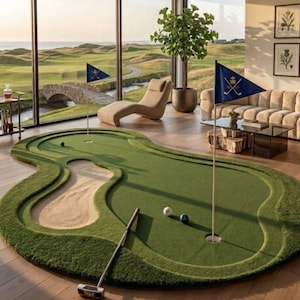 May include: An indoor putting green mat featuring a sand trap, two holes with blue flags displaying a gold crossed-club emblem, a putter, and two golf balls. The green is shaped with curved edges and sits on a wooden floor in a room.
