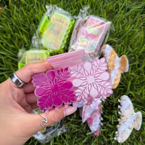 May include: A collection of hair claw clips in various colors and patterns, including pink, white, and gold. The clips feature floral designs and are displayed on a bed of green grass. Some clips are packaged in clear plastic.
