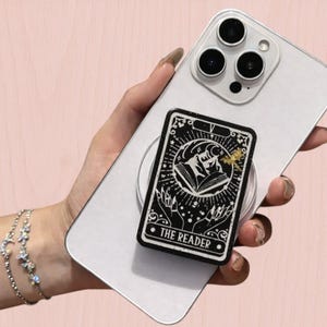 May include: A black and white phone grip featuring a tarot card design with the text "THE READER". The grip is attached to a silver smartphone. The design includes an illustration of a figure reading a book, surrounded by stars and lines.