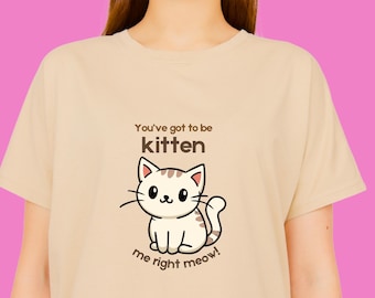 Puede incluir: Camiseta beige con una ilustración de un gato de dibujos animados y el texto "You've got to be kitten me right meow!". El diseño incluye un gato blanco con bigotes negros y una cola a rayas. La camiseta es de color neutro.