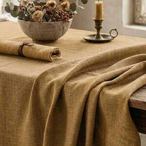 May include: A warm-toned, textured linen tablecloth draped over a wooden table. A rolled napkin with a leather strap, a stone bowl with dried flowers, and a brass candle holder with a lit candle are on the table. The scene evokes a rustic, cozy atmosphere.