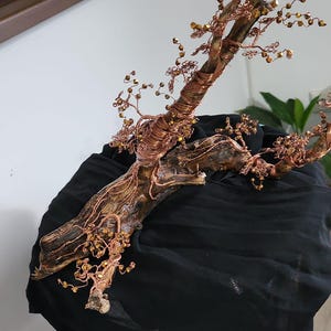 Puede incluir: Escultura artesanal de árbol de alambre de cobre con base de madera natural. El árbol presenta intrincadas ramas de alambre y pequeños acentos de cuentas doradas, creando una pieza decorativa única. La escultura se exhibe sobre tela negra.