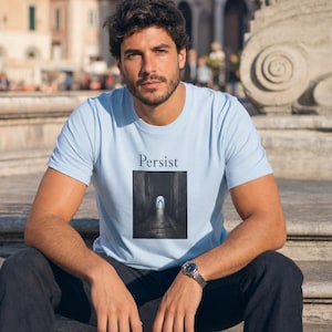 May include: A light blue t-shirt with the word "Persist" above a black and white graphic of a tunnel. The graphic shows a long, dark tunnel with a light at the end. The shirt is made of a soft material.