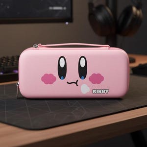 May include: Pink Nintendo Switch carrying case featuring a cartoon face with large eyes, pink cheeks, and a small mouth. The word "KIRBY" is printed below the face. The case has a zipper closure and a small handle.