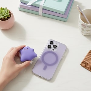 May include: A purple Ditto-shaped phone grip beside a clear phone case with a purple magnetic ring. A small succulent in a pink pot, a stack of notebooks, and a floral mug are on the white surface.