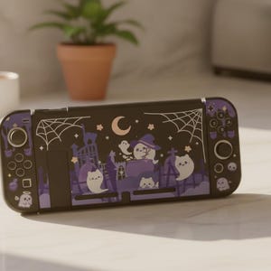 May include: A handheld gaming console featuring a black and purple skin with a graphic illustration of ghosts, a crescent moon, stars, and spiderwebs. The design includes small white ghost figures and a purple castle silhouette on the front panel.