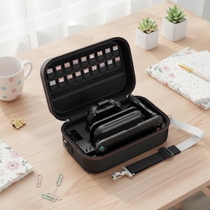 May include: Black carrying case with a red trim, open to reveal a gaming console, controllers, and game cartridges. The case has a shoulder strap and is on a wooden surface with a mug, paper, and small plants.