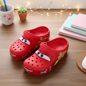 Könnte beinhalten: Ein Paar leuchtend rote Kinderclogs mit einem Cartoon-Auto-Design, einschließlich blauer Augen und einem lächelnden Mund. Die Schuhe haben Belüftungslöcher und eine gelb-orange Grafik. Die Clogs liegen auf einer Holzoberfläche.
