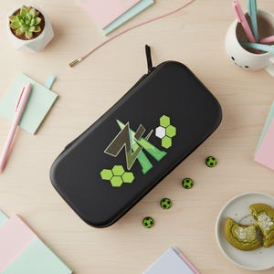 May include: Black rectangular case with a green and white graphic design and a zipper. Small soccer ball decorations are scattered around the case. The case is on a light-coloured wooden surface. The case is likely for a games console.