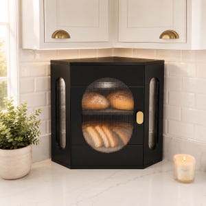 May include: A black corner bread box with a triangular shape, featuring a central oval door with ribbed glass and two vertical side panels of ribbed glass. The unit includes a gold-toned oval handle and two internal shelves holding bread.