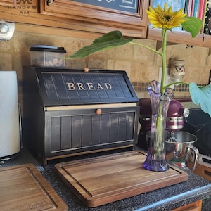 May include: A black wooden bread box with a hinged lid featuring the word BREAD in light-colored lettering, accompanied by a rectangular wooden cutting board and a purple glass vase containing a single yellow sunflower on a kitchen counter.
