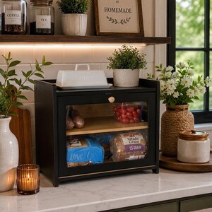 May include: A black bread box with a clear glass front door and a wooden shelf inside. The box contains a loaf of bread and a container of cherry tomatoes. A white butter dish sits on top of the box, which is placed on a white countertop.