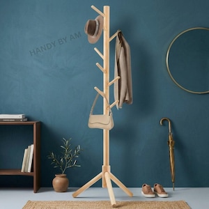 May include: A light-colored wooden coat rack with multiple pegs for hanging items. A brown hat, beige purse, and tan jacket hang on the rack. The rack has a tripod base and is in a room with a blue wall.