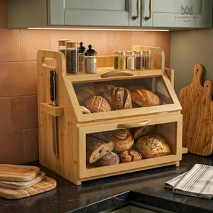 May include: A two-tier bamboo bread box with clear glass front panels and a side knife slot. The unit holds various loaves of bread and is topped with a shelf containing several small glass spice jars. The wood has a natural light brown finish.