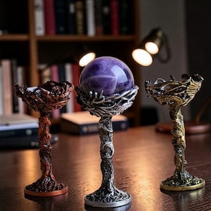 May include: Three decorative crystal ball holders. One holds a purple crystal ball. The holders are made of metal with intricate designs. The holders are in shades of bronze, silver, and gold. The holders are on a wooden surface.