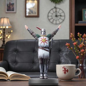May include: A figurine of a knight in silver armor with arms raised, featuring a sun emblem on the chest. The knight stands on a gray base, with a sword at its side. A coffee cup with a red cross design sits nearby.
