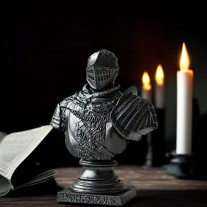 May include: A dark gray bust of a knight in full armor, including a helmet and shoulder guard. The bust is mounted on a pedestal with the words "DARK SOULS" at the base. The background includes candles and an open book.