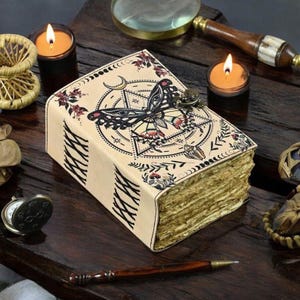 Puede incluir: Un diario de cuero beige con un diseño de mariposa y celestial. La portada presenta una ilustración detallada de una mariposa, una luna creciente y otros símbolos. Las páginas tienen un borde dorado. También se ven dos velas y un bolígrafo.