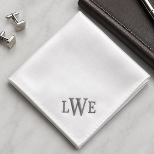 Puede incluir: Un pañuelo de bolsillo cuadrado de color blanco con un acabado suave similar al satén y un monograma centrado bordado en hilo gris con las letras LWE. La tela está doblada en forma de diamante, mostrando bordes cosidos limpios.