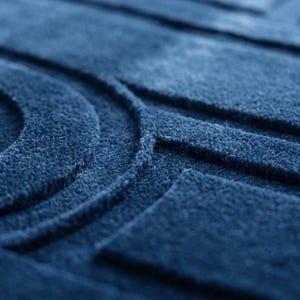 K&ouml;nnte beinhalten: Ein tiefblauer Teppich mit einer strukturierten Oberfl&auml;che und erhabenen geometrischen Mustern. Das Design besteht aus konzentrischen Kreisformen und geraden rechteckigen Linien, die einen taktilen, dreidimensionalen Effekt erzeugen.