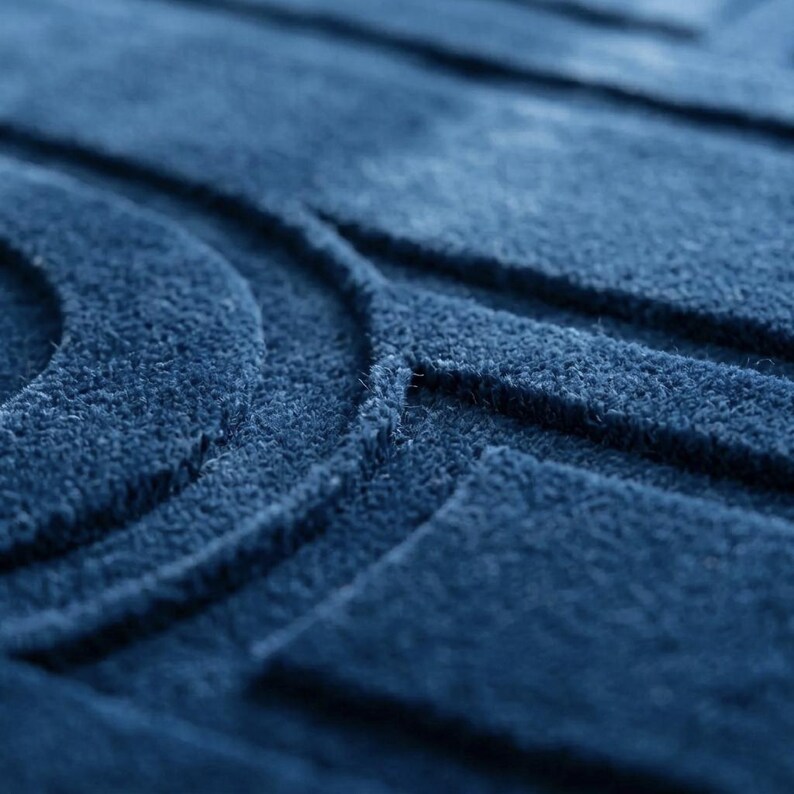 K&ouml;nnte beinhalten: Ein tiefblauer Teppich mit einer strukturierten Oberfl&auml;che und erhabenen geometrischen Mustern. Das Design besteht aus konzentrischen Kreisformen und geraden rechteckigen Linien, die einen taktilen, dreidimensionalen Effekt erzeugen.