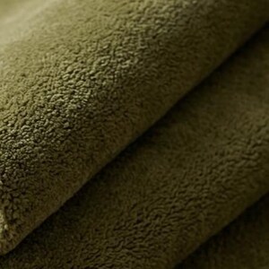 May include: A close-up view of olive green fabric with a dense, soft, and fuzzy texture. The material is folded over itself, creating visible layers and shadows that highlight the thick, plush pile of the textile surface in a solid color.