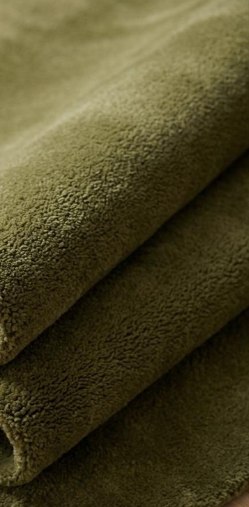 May include: A close-up view of olive green fabric with a dense, soft, and fuzzy texture. The material is folded over itself, creating visible layers and shadows that highlight the thick, plush pile of the textile surface in a solid color.
