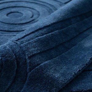 K&ouml;nnte beinhalten: Eine tiefblaue Badematte mit einer weichen, strukturierten Oberfl&auml;che, die mit erhabenen konzentrischen Kreisen und geraden parallelen Linien versehen ist. Der Stoff hat einen dichten Flor, der ein subtiles Licht- und Schattenspiel erzeugt.