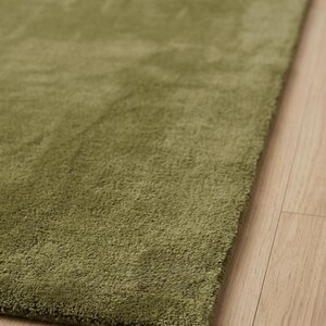 May include: A rectangular area rug featuring a solid olive green color with a dense, short-pile texture. The rug is positioned at an angle, showcasing its uniform surface and clean, straight edges against a light-toned wooden floor surface.