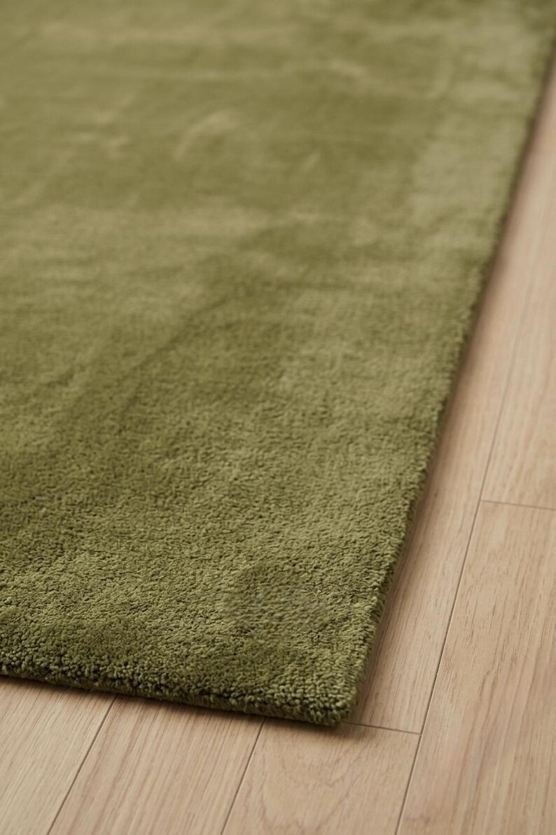 May include: A rectangular area rug featuring a solid olive green color with a dense, short-pile texture. The rug is positioned at an angle, showcasing its uniform surface and clean, straight edges against a light-toned wooden floor surface.