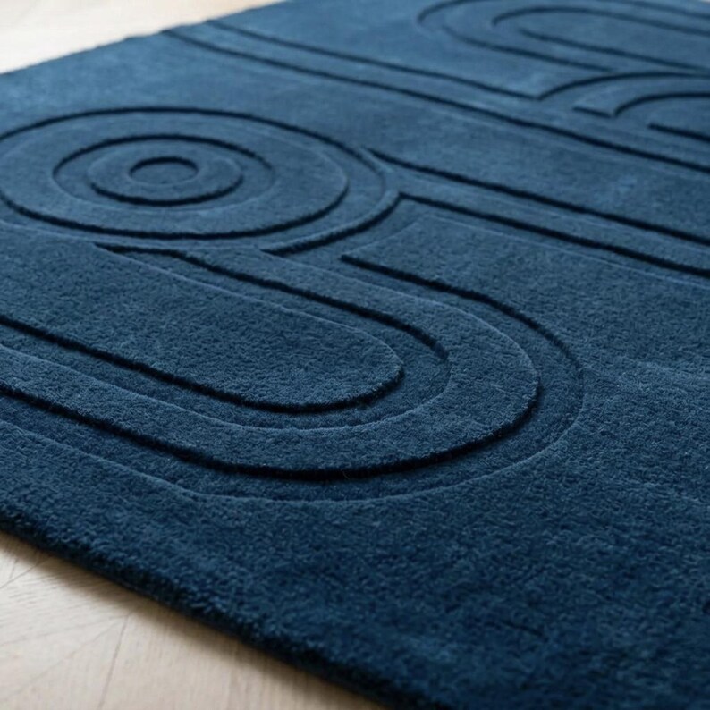 K&ouml;nnte beinhalten: Ein tief dunkelblauer Teppich mit einer erhabenen, geschnitzten Textur, die eine Reihe konzentrischer Kreise und l&auml;nglicher, abgerundeter geometrischer Linien auf der Oberfl&auml;che bildet und ein haptisches, dreidimensionales Muster erzeugt.